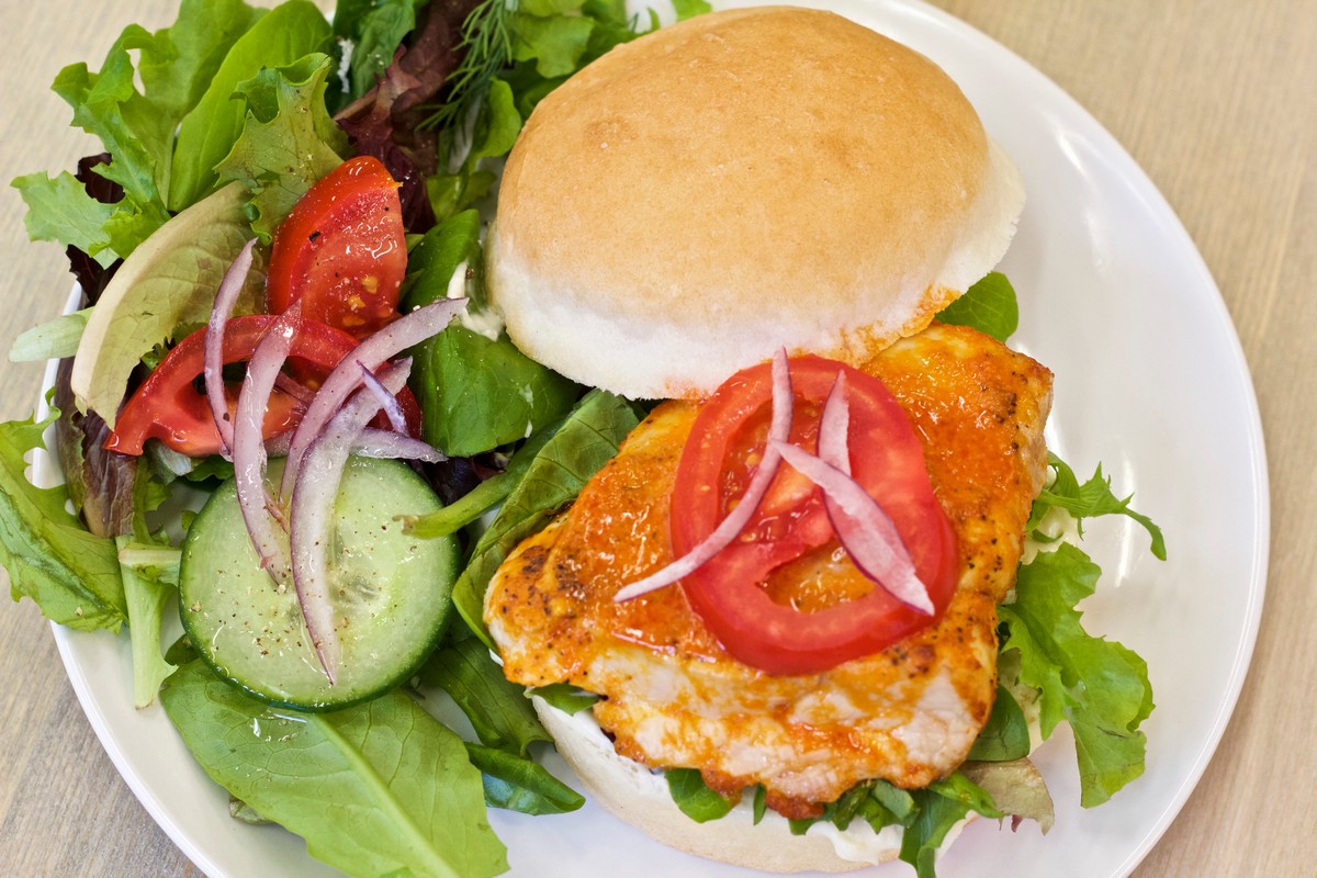 Buffalo Chicken on gluten free bun surrounded by salad on a white plate on wooden board