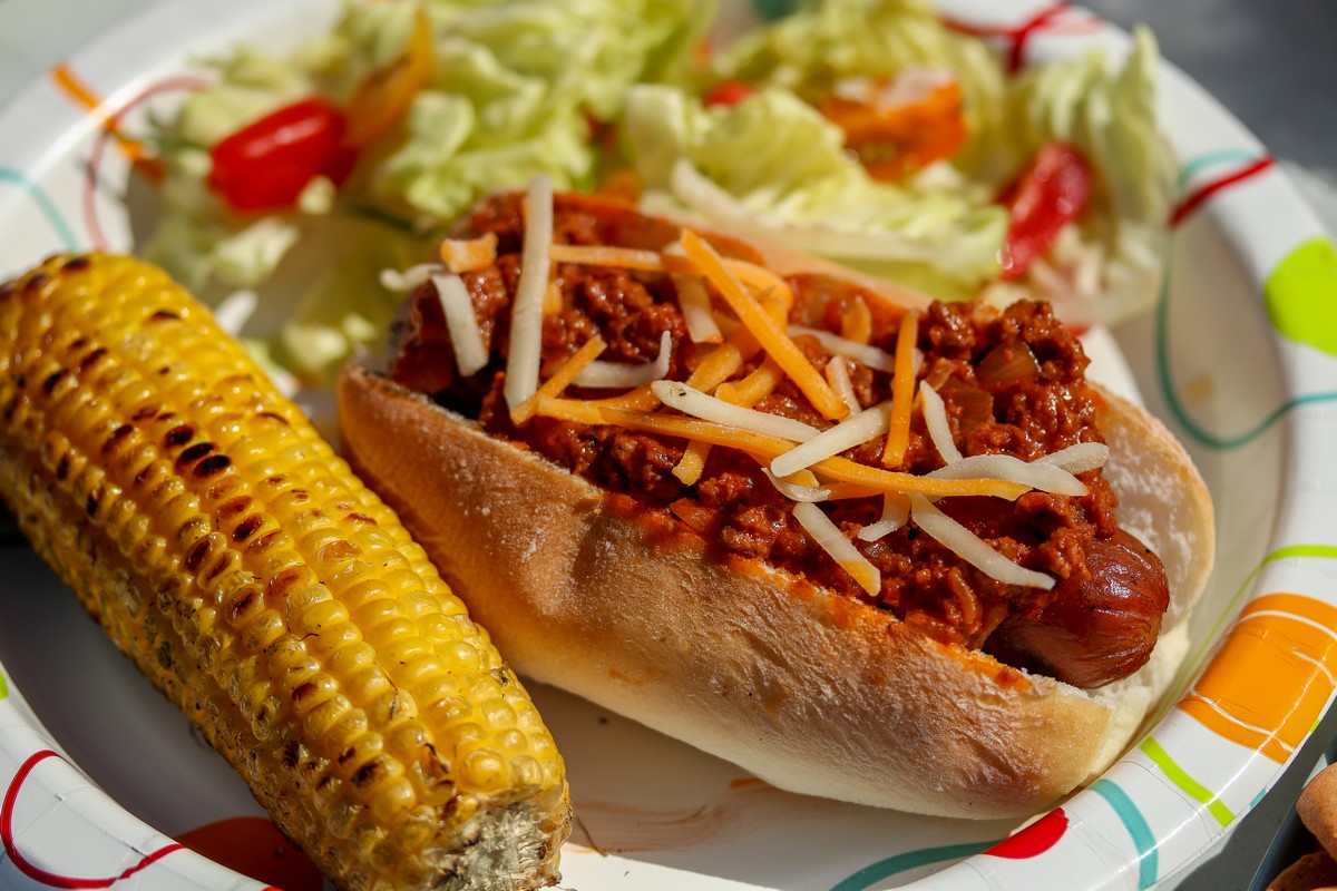 Gluten-free chili dog on white paper plate surrounded by corn cob & salad
