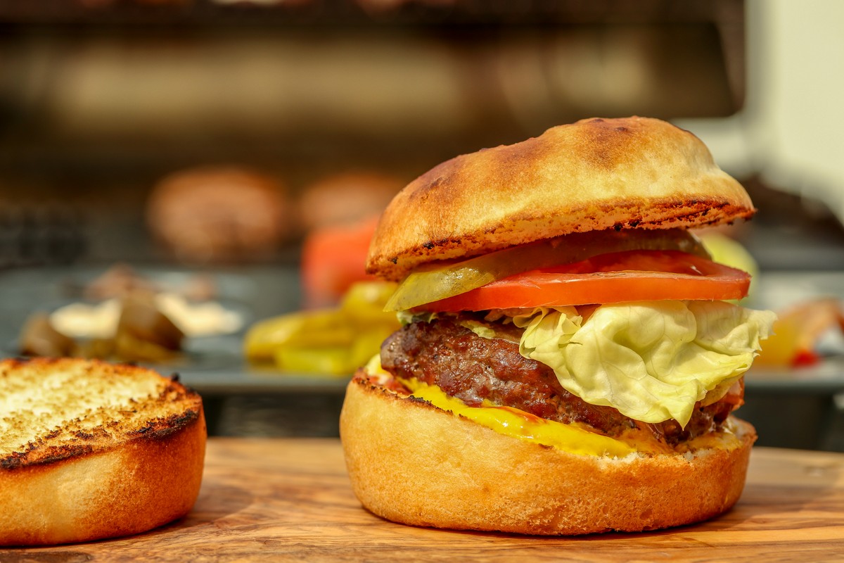 Gluten-free bun with beef patty on wooden board surrounded by condiments with bbq in background