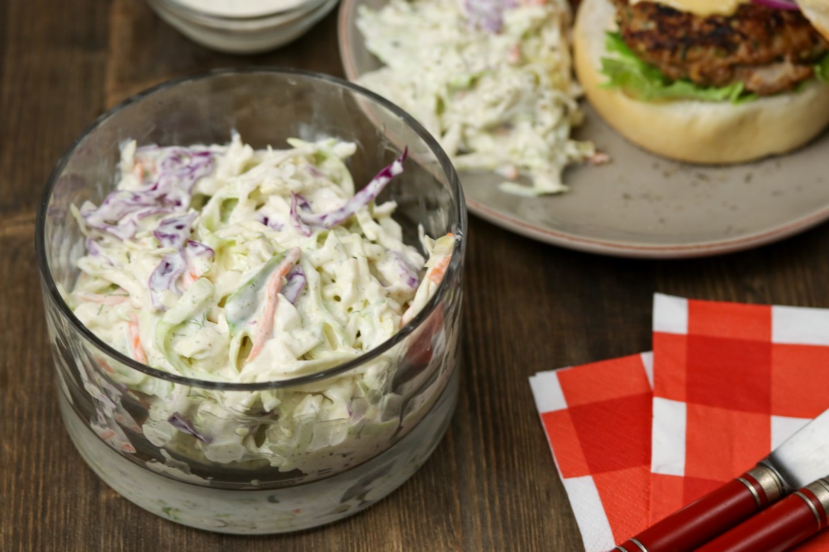 Creamy coleslaw in a glass dish on wooden board, burger in background
