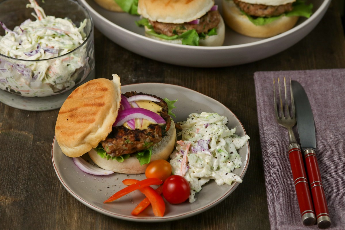 Gluten-free Hamburger Bun filled with Herbed Turkey patty, red onions on a grey plate, wooden table with red cutlery and a full pan of burgers behind