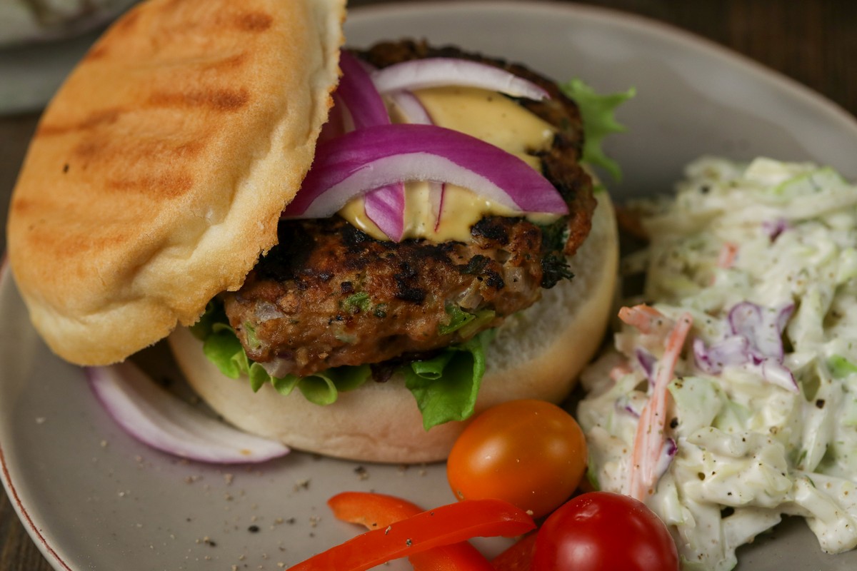 Gluten-free Hamburger Bun filled with Herbed Turkey patty, red onions on a grey plate, wooden table with red cutlery and a full pan of burgers behind