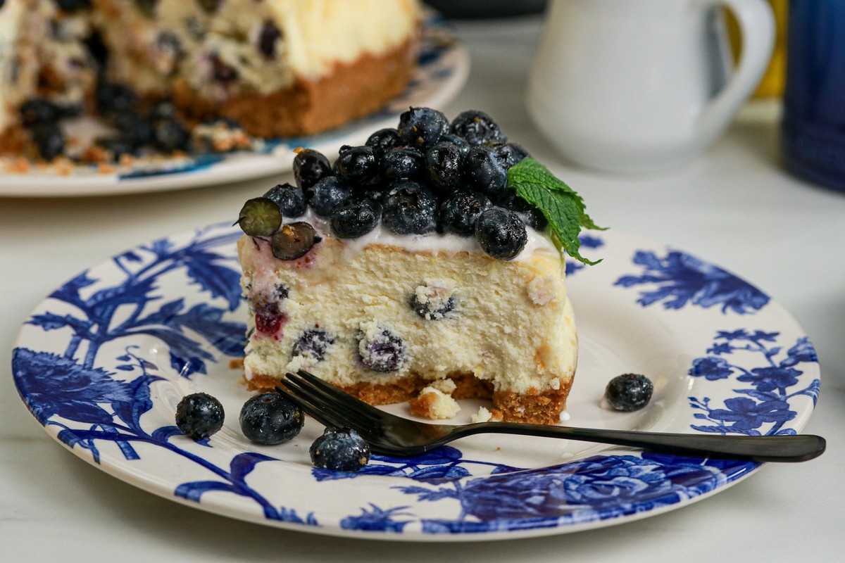 Gluten-free Lemon Blueberry Cheesecake slice on blue white plate surrounded by lemon various cups and cutlery