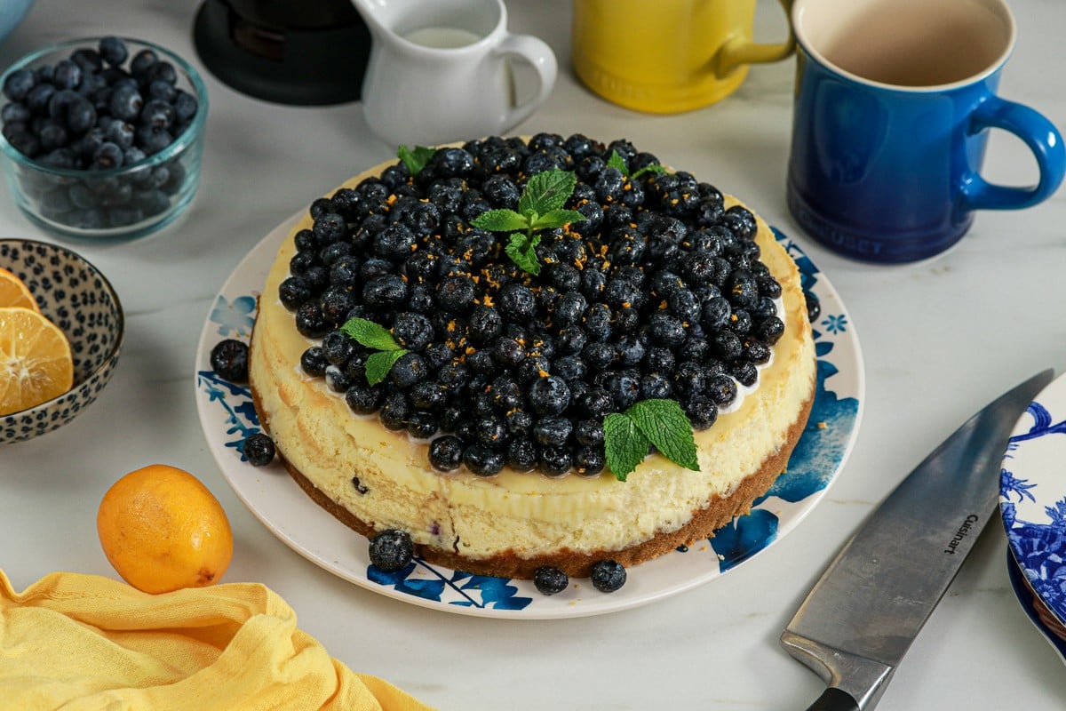 Gluten-free Lemon Blueberry Cheesecake on blue white plate surrounded by lemon various cups and cutlery