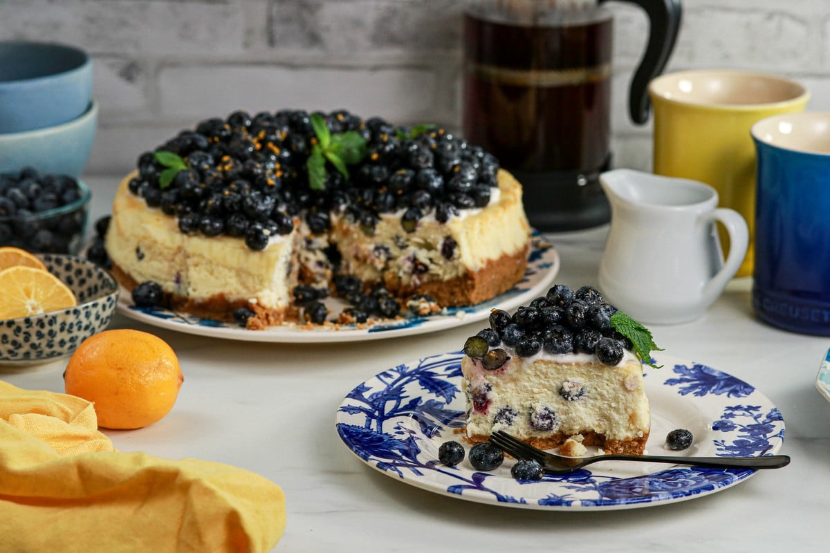 Gluten-free Lemon Blueberry Cheesecake sliced on blue white plate surrounded by lemon various cups and cutlery