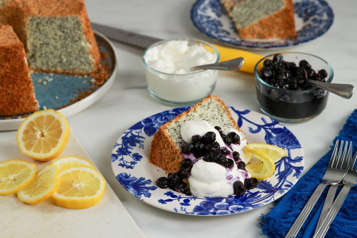 Gluten-free Poppy Seed Chiffon Cake on a blue white plate covered in cream & blueberry compote with lemons and glass bowls